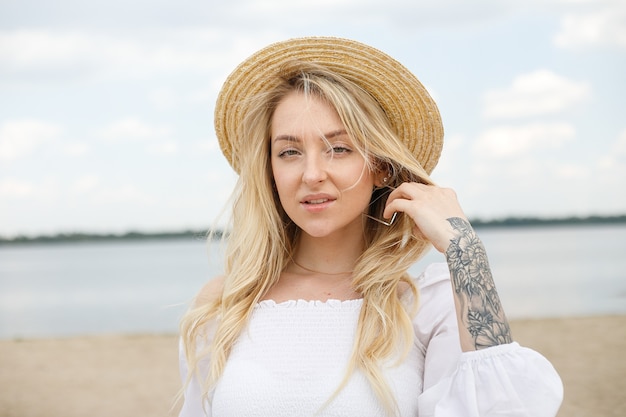 Incroyable jeune fille aux cheveux blonds bouclés se développant du vent avec le chapeau de paille sur la tête dans le haut blanc contre la mer, sourit doucement