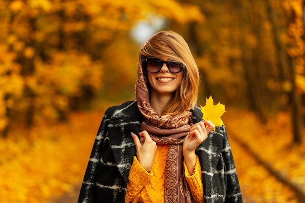 Incroyable portrait d'une femme heureuse avec un sourire de beauté en manteau de mode, pull tricoté, écharpe et lunettes de soleil vintage se promène dans la nature avec de jolies feuilles d'automne jaunes. Style féminin à l'extérieur