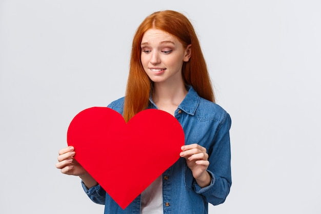 Indécise, mignonne et timide, une femme rousse timide veut se confesser en amour, montrer de la sympathie incertaine donner un cadeau le jour de la Saint-Valentin ou non, tenant un grand coeur rouge, une lèvre mordante inquiète