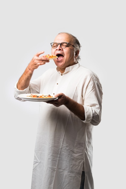 Indien asiatique senior ou vieil homme mangeant de la pizza en se tenant isolé contre le mur blanc
