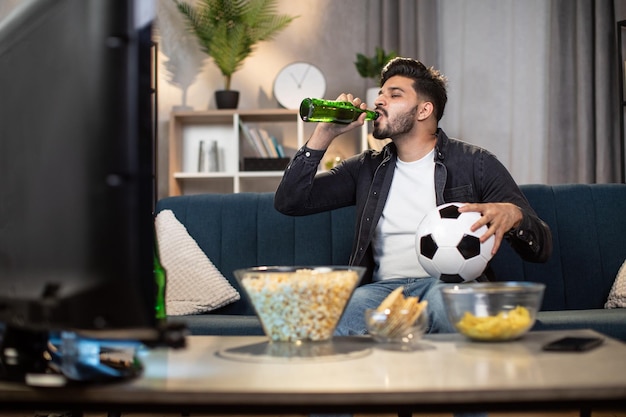 Un Indien qui boit de la bière en regardant un match de football à la maison