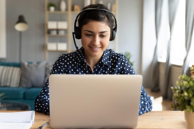 Une Indienne souriante du millénaire dans un casque sans fil est assise au bureau dans le salon et étudie en ligne sur un ordinateur portable.