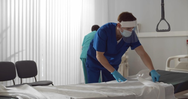 Infirmier en masque de sécurité uniforme bleu et gants changeant les draps du lit d'hôpital dans la salle