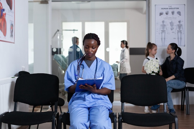 Infirmière afro-américaine remplissant les documents du patient assis sur les chaises de réception de la tour médicale. Jeune médecin rédigeant des ordonnances dans la salle d'attente de l'hôpital. Membre du personnel médical examinant la liste des patients