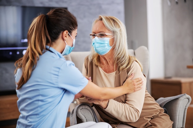 Infirmière assise à la maison avec un retraité et la réconfortant pendant la pandémie de coronavirus. Ils ont tous les deux des masques faciaux.