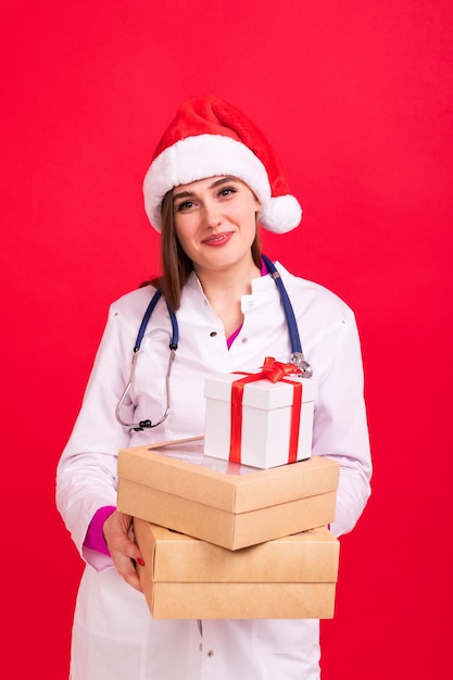 Une infirmière dans un chapeau de Père Noël tient des boîtes avec des cadeaux sur fond rouge Félicitations du médecin aux patients