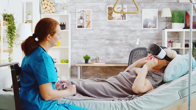 Infirmière dans une maison de retraite donnant à une vieille dame handicapée un casque VR pour soulager la douleur et la distraire pendant qu'elle est allongée dans un lit d'hôpital