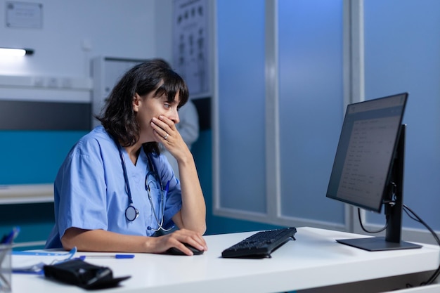 Infirmière fatiguée utilisant l'ordinateur sur le bureau en s'endormant, travaillant tard. Assistant médical regardant l'écran du moniteur et se sentant somnolent après des heures supplémentaires la nuit dans une armoire de soins de santé.