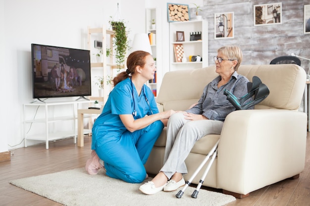 Infirmière à genoux parlant avec une vieille femme dans une maison de retraite. Vieille femme avec des béquilles.