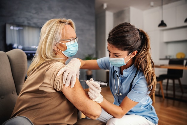 Infirmière avec masque assis à la maison et administrant une injection à une vieille femme pendant une épidémie de corona.
