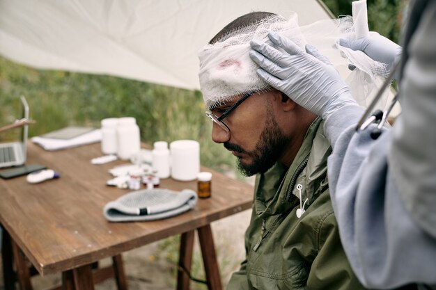 Infirmière méconnaissable dans des gants mettant un bandage sur la tête blessée d'un réfugié dans le camp