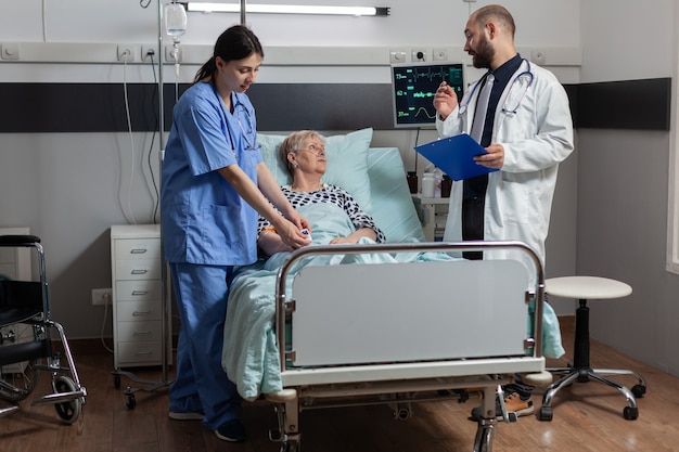 Infirmière médicale attachant un oxymètre sur une patiente âgée, allongée sur un lit d'hôpital discutant avec un médecin du diagnostic et du traitement pendant qu'elle respire avec l'aide d'un masque oxygem
