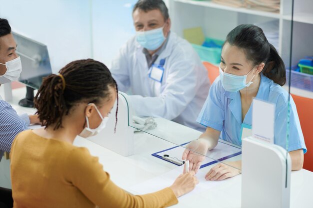 Infirmière médicale derrière un écran de verre aidant le patient dans un masque de protection à remplir le formulaire à la réception de l'hôpital.