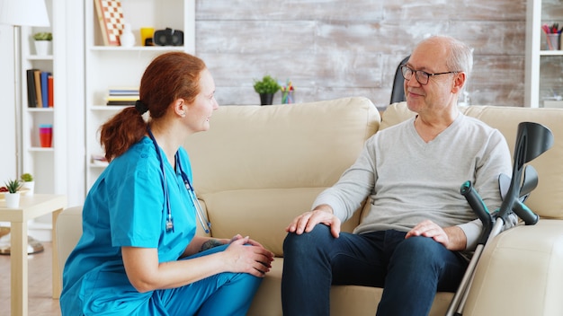 Infirmière parlant et prenant soin d'un vieil homme handicapé assis sur le canapé dans une maison de soins infirmiers. Travailleur social et aide-soignant