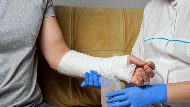 Une infirmière portant des gants médicaux utilise des ciseaux pour couper le pansement de gypse du bras guéri d'un jeune homme en T-shirt gris, assis sur un canapé dans le salon.