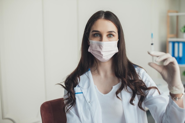 Infirmière posant avec une seringue pour l'injection de vaccin portant des gants en caoutchouc de robe blanche et un masque médical co...