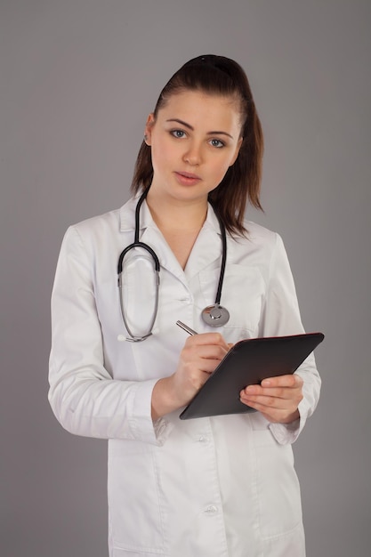 Infirmière en robe blanche et avec stéthoscope écrit quelque chose sur fond gris