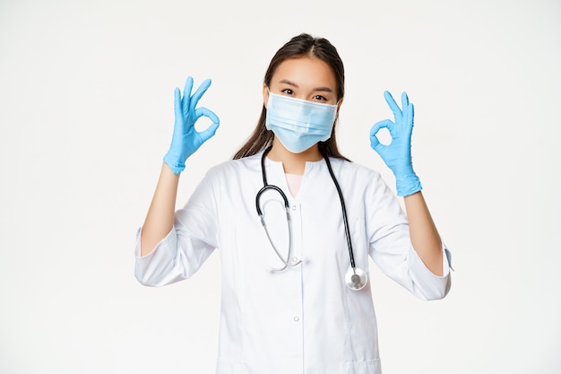 Infirmière souriante, femme médecin asiatique portant des gants en caoutchouc, un masque médical montre des signes ok ok se dresse contre ...