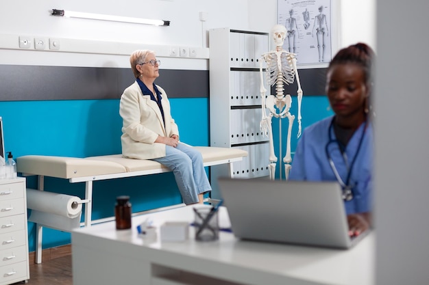 Photo infirmière spécialisée travaillant dans le traitement des soins de santé en tapant l'expertise de la maladie sur ordinateur tandis qu'une patiente âgée debout sur son lit attend le diagnostic de la maladie dans le bureau de l'hôpital. aide au soutien