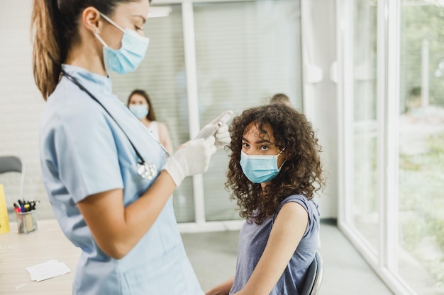 Infirmière tenant une seringue et faisant une dose d'injection de vaccination Covid-19 dans l'épaule d'une patiente adolescente portant un masque.