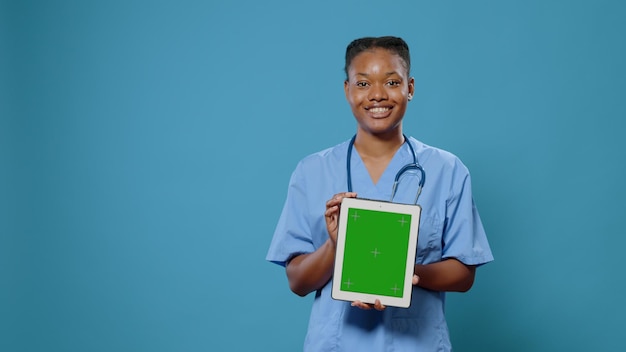 Infirmière tenant verticalement une tablette avec écran vert en studio. Assistant médical en uniforme, utilisant une clé chroma avec un modèle de maquette isolé et un arrière-plan sur l'affichage du gadget