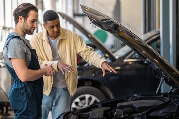 Photo une information important. jeune mécanicien caucasien barbu regardant attentivement le capot de la voiture et le client à la peau foncée racontant le problème debout dans l'atelier