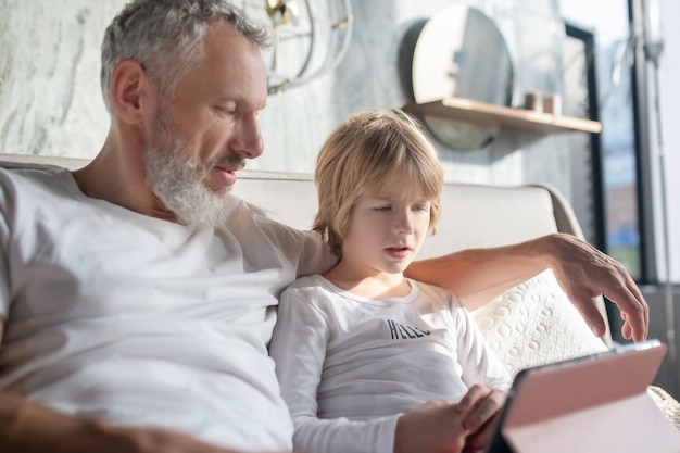 Informations utiles. Papa aux cheveux gris attentionné en tshirt blanc étreignant l'enfant attentif à la tablette sur le lit