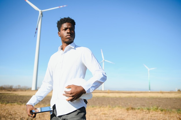 Ingénieur africain portant un casque blanc debout avec une tablette numérique contre une éolienne