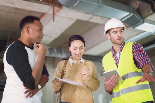 ingénieur architecte utilisant une tablette tout en montrant des plans de conception de maison à un jeune couple multiethnique sur un chantier de construction