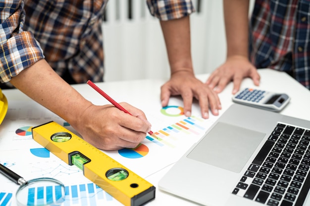 L'ingénieur asiatique et l'équipe partenaire apprennent et parlent avec le graphique de plan de projet de plan de maison et le casque jaune et les outils de construction d'ingénierie sur le travail de table