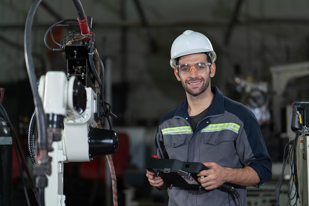 Ingénieur en automatisation mâle souriant tenir la carte à distance pour contrôler la machine à souder à bras robotique en usine