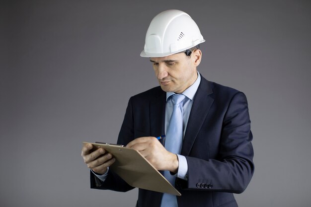 Ingénieur en chef dans un casque prend des notes sur le presse-papiers isolé sur mur gris