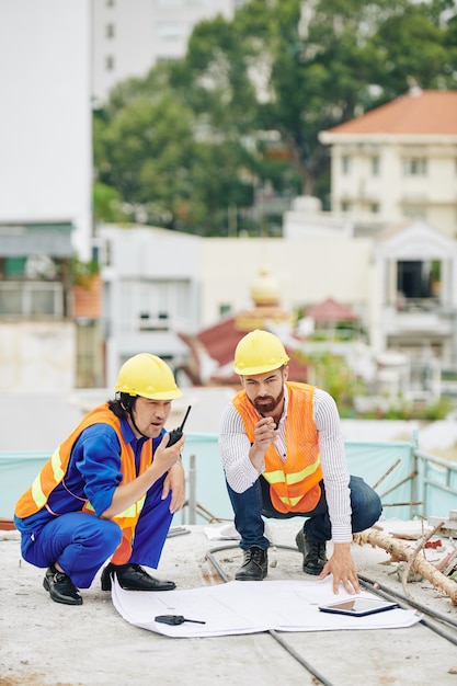 Ingénieur en chef vérifiant le plan de construction lorsque l'entrepreneur contrôle le travail des constructeurs via talkie-walkie