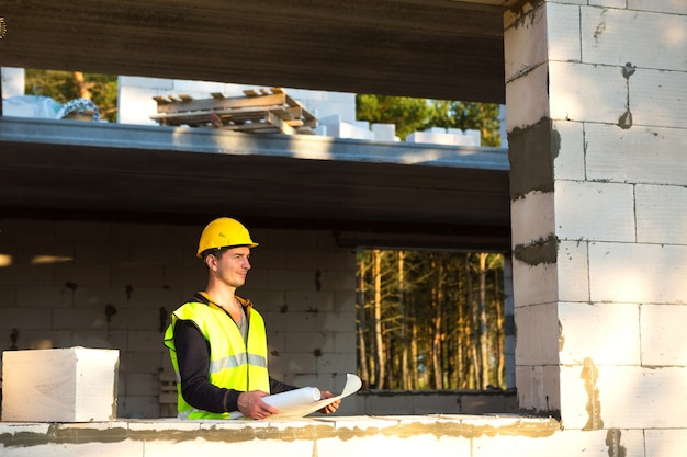 Un ingénieur en construction et designer portant un casque jaune étudie un dessin d'un bâtiment sur un chantier de construction. Une maison faite de blocs de béton poreux, un ouvrier dans le processus à la fenêtre et au mur