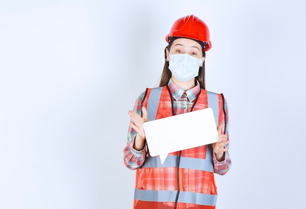 Ingénieur en construction femelle dans un masque de sécurité et un casque rouge tenant un panneau d'information vierge rectangle.