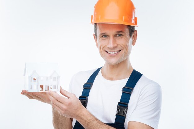 Ingénieur en construction. Joyeux homme gentil positif tenant un modèle de maison et souriant tout en travaillant comme ingénieur en bâtiment