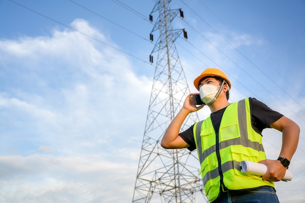 Ingénieur électricien asiatique tenant un plan à la centrale électrique en activité et envisage d'inspecter les poteaux haute tension de la centrale électrique