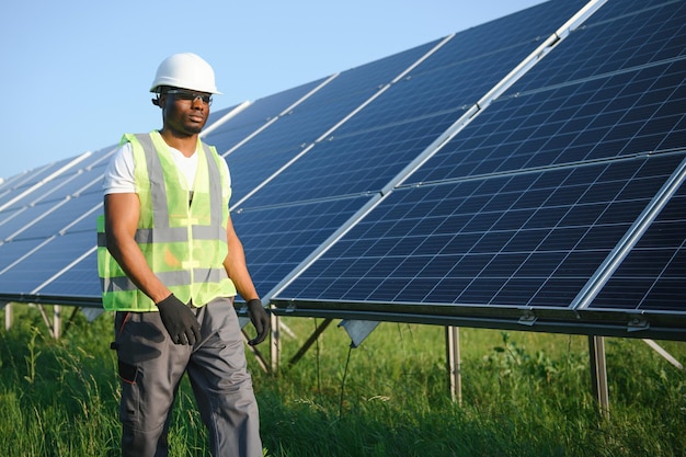 Ingénieur en énergie compétent en salopette grise et casque orange vérifiant les panneaux solaires tout en marchant sur le terrain Homme afro-américain transportant un presse-papiers et un conteneur avec des instruments