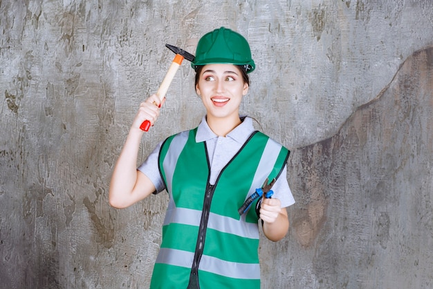 Ingénieur féminin en casque vert tenant des pinces et une hache à manche en bois pour un travail de réparation