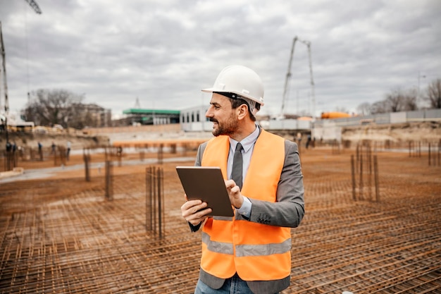 Un ingénieur heureux vérifie les travaux de construction sur une tablette tout en se tenant sur le chantier