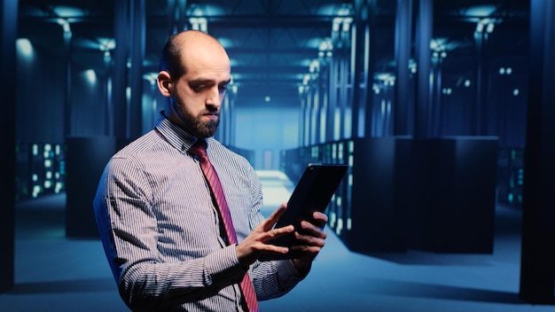 Ingénieur informatique du centre de données debout dans la salle des serveurs avec des armoires rack, inspectant le service de données volumineuses de cloud computing. Administrateur système exécutant la sauvegarde réseau sur tablette, intelligence artificielle.