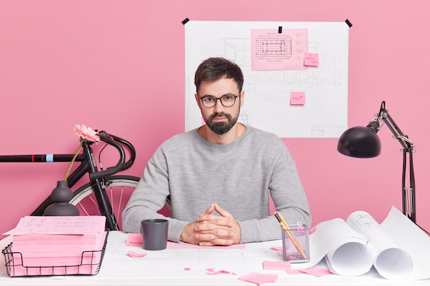 Un ingénieur masculin professionnel barbu et confiant, prêt à vous aider dans votre nouvelle maison, pose un projet de conception au bureau et boit du café entouré de bluprints fait du travail sur papier travaille au bureau à domicile