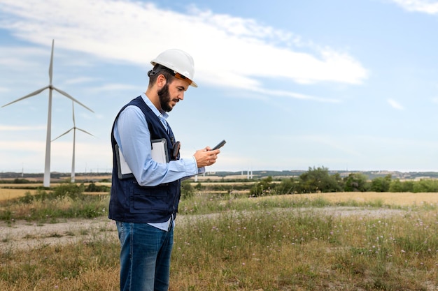 Ingénieur portant un casque et une veste vérifiant les données sur le téléphone inspectant une ferme d'éoliennes Copier l'espace