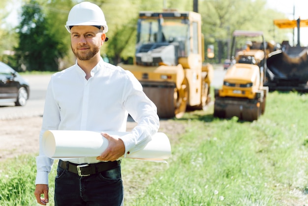 Ingénieur souriant avec casque debout devant une pelle sur le chantier de construction de routes