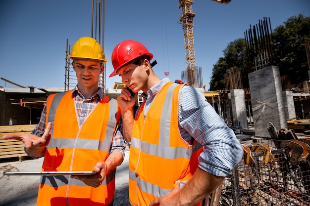 Photo l'ingénieur en structure et l'architecte vêtus de gilets de travail orange et de chauves-souris dures discutent du processus de construction par téléphone et utilisent une tablette sur le chantier ouvert près de la grue. .