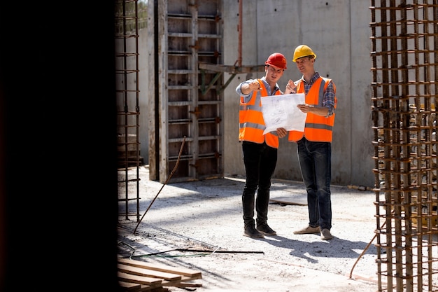 L'ingénieur en structure et le directeur de la construction portant des gilets de travail orange et des casques durs explorent la documentation de construction sur le chantier près des cadres en acier. .