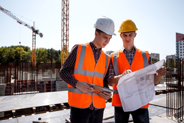 L'ingénieur en structure et le directeur de la construction vêtus de gilets de travail orange et de casques discutent de la documentation sur le chantier en plein air à côté de la grue .