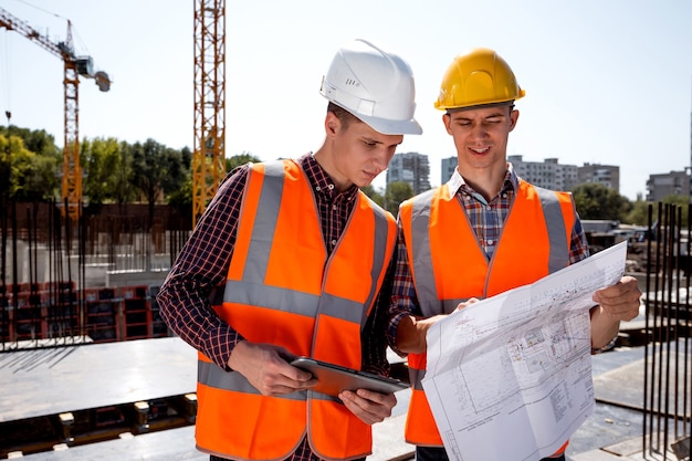 L'ingénieur en structure et le directeur de la construction vêtus de gilets de travail orange et de casques discutent de la documentation sur le chantier en plein air à côté de la grue .