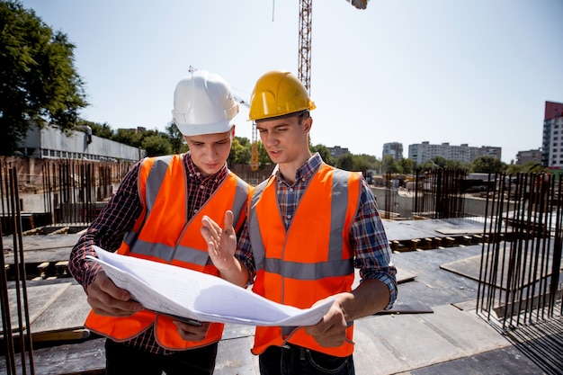 L'ingénieur en structure et le directeur de la construction vêtus de gilets de travail orange et de casques discutent de la documentation sur le chantier en plein air à côté de la grue .