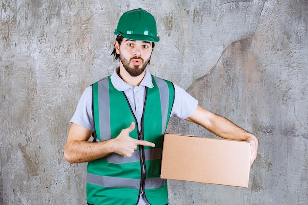 Ingénieur en tenue jaune et casque tenant une boîte en carton et a l'air confus et réfléchi.